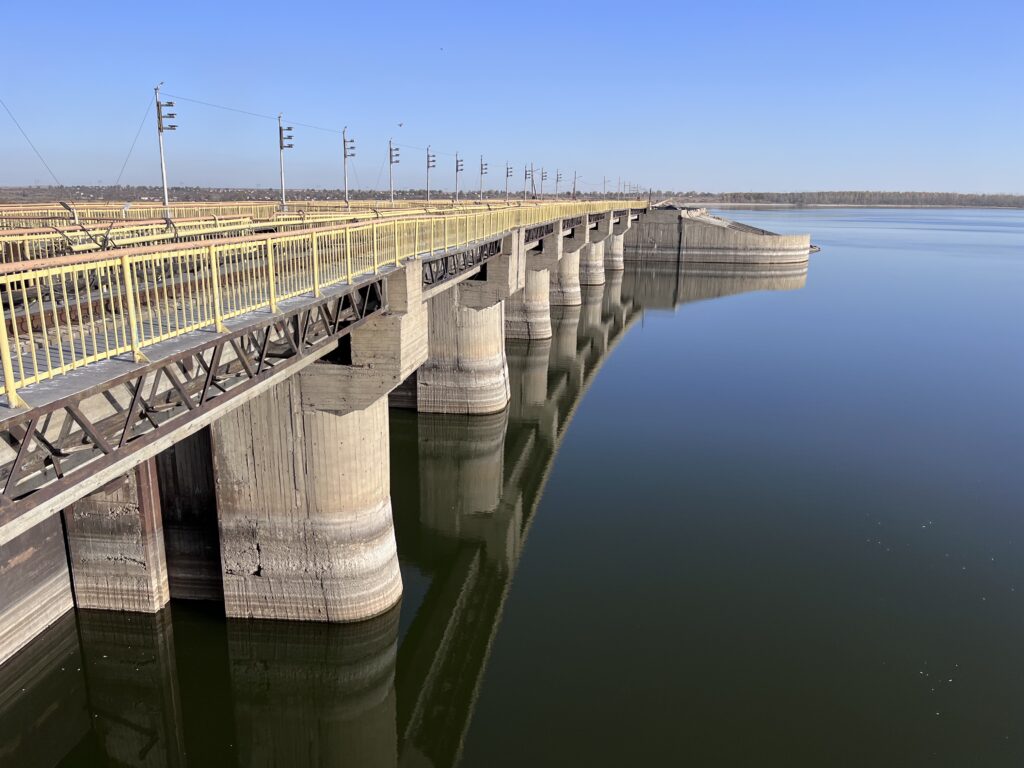 Concrete dam wall with yellow railings and metal trusses extending over the water, supported by cylindrical pillars, with water levels clearly visible on the concrete and calm reservoir water alongside.