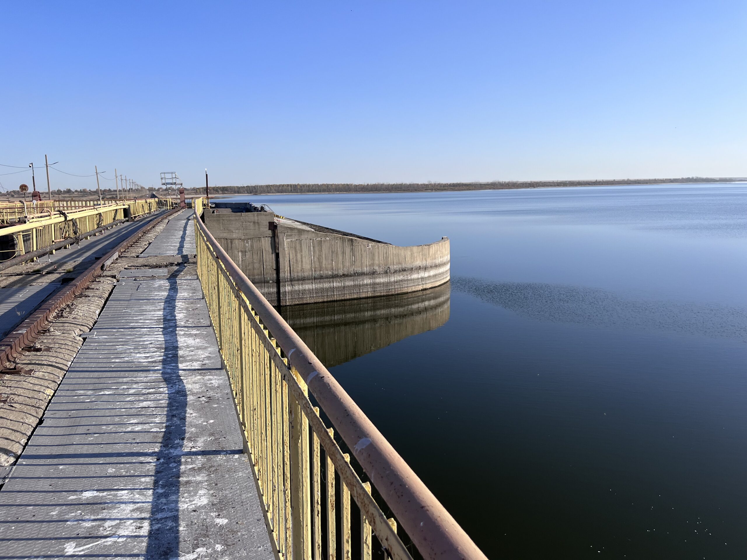 Concrete dam structure with a long walkway and yellow railings extending across multiple piers above calm reservoir water, with reflections visible below.