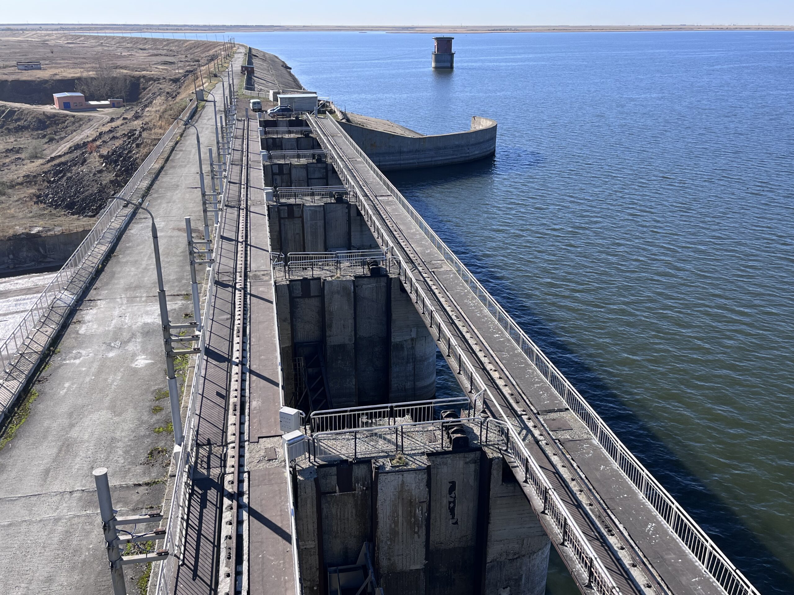 Elevated view along a concrete dam with multiple vertical sluice gates and metal railings, bordered by a service road on the left and a large water reservoir on the right.