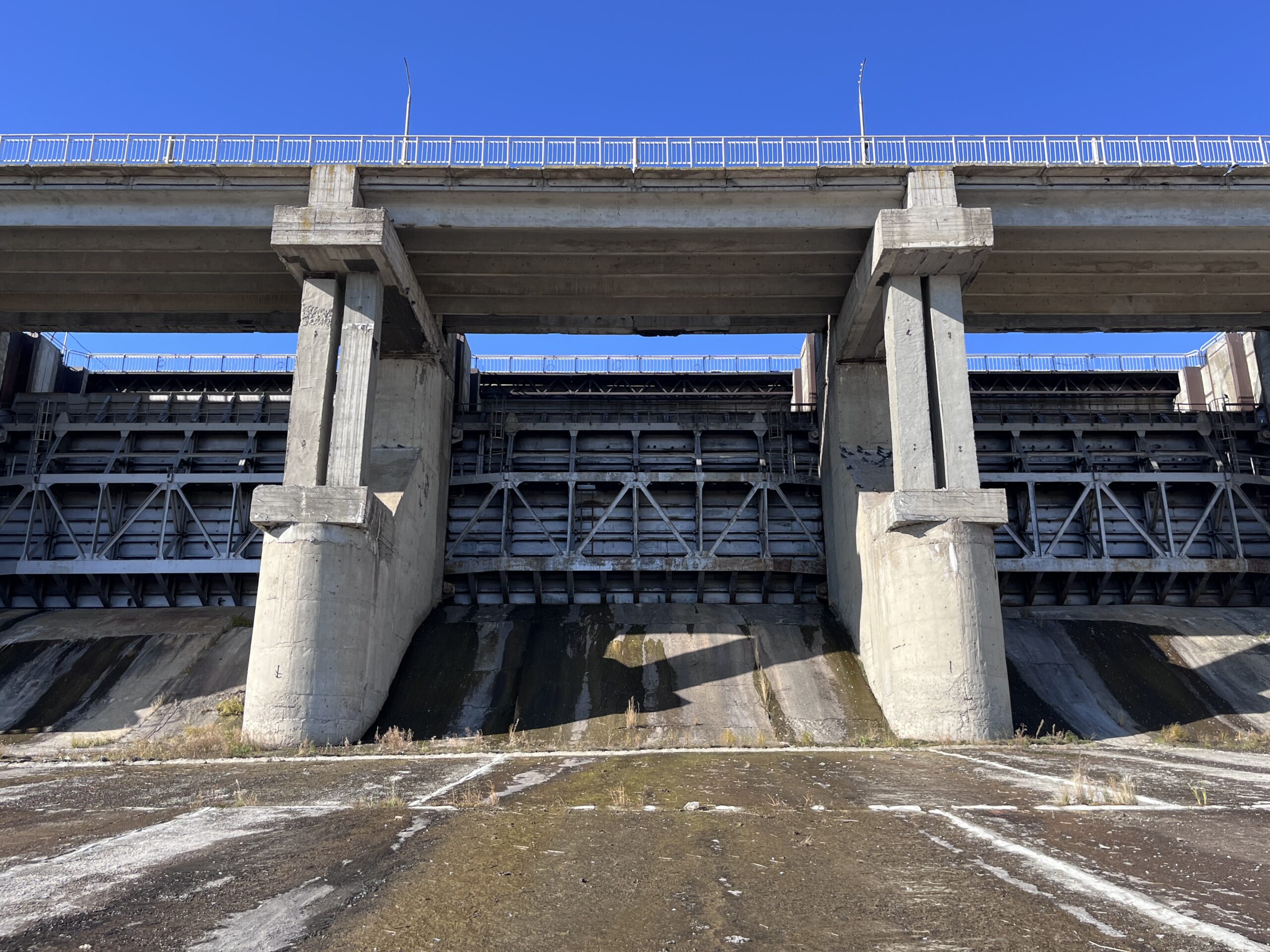 Concrete dam with gated spillways viewed from downstream, with low water levels exposing the spillway surface and shallow pooled water in the foreground.