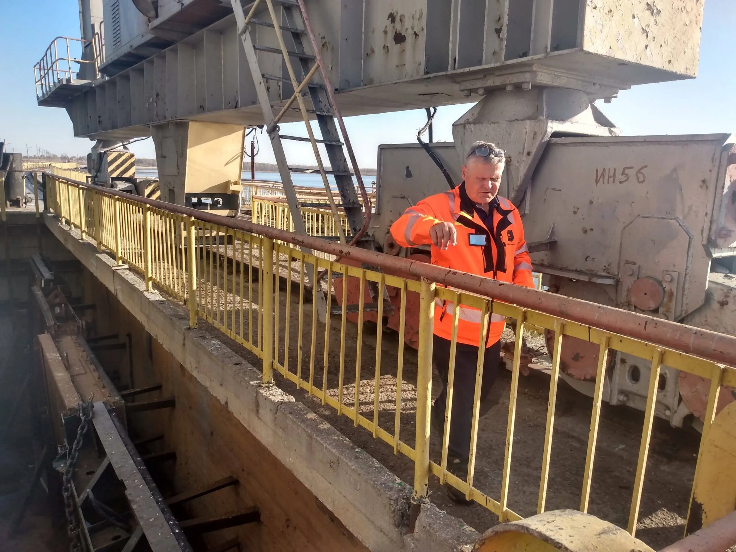 Engineer inspecting a large dam gate mechanism from a walkway with yellow railings, with water visible in the background.