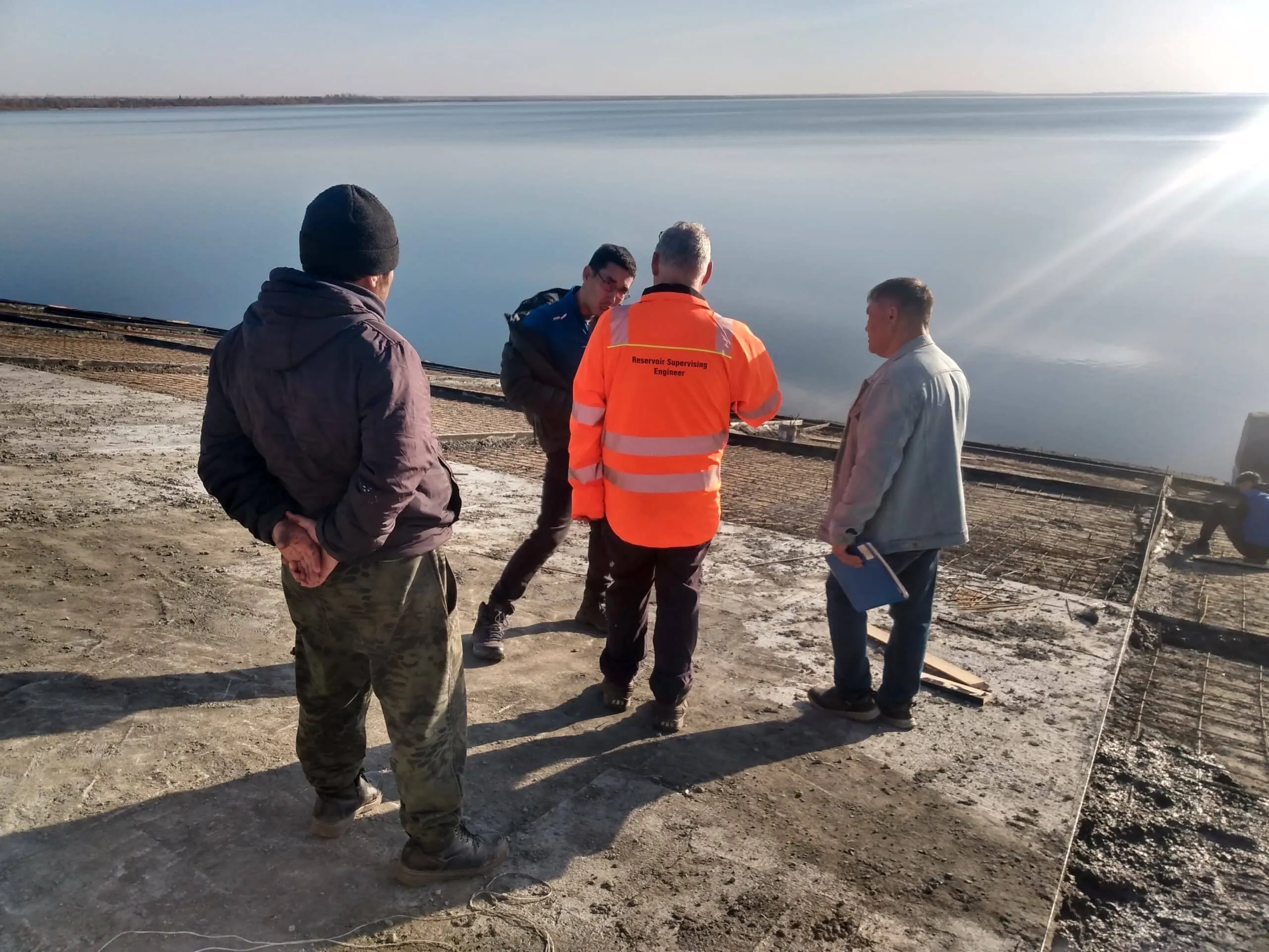 Group of engineers and site staff standing on a concrete dam structure beside a reservoir, discussing inspection works near exposed reinforcement and construction areas.