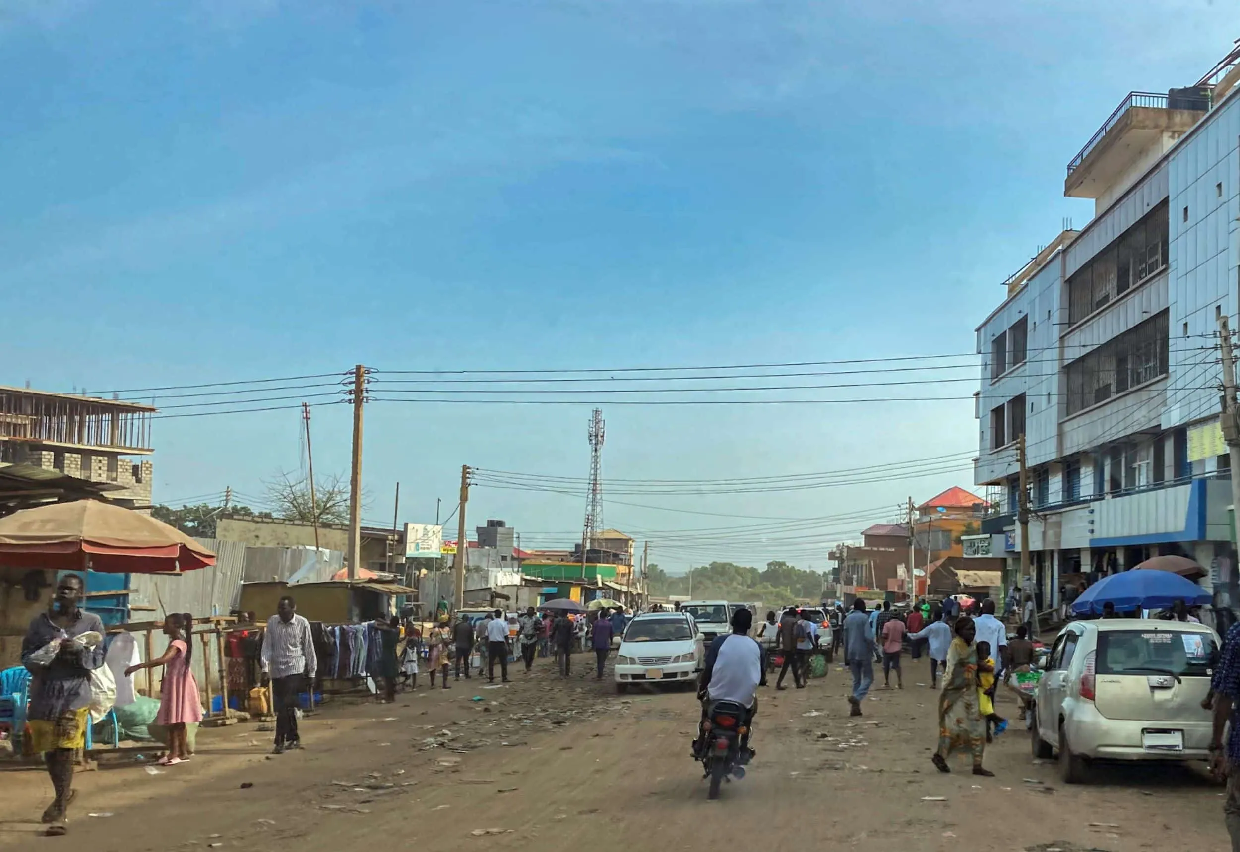 Busy street scene (Juba, South Sudan) with pedestrians, cars, and market stalls along a wide dirt road, lined with low-rise buildings and overhead power lines.