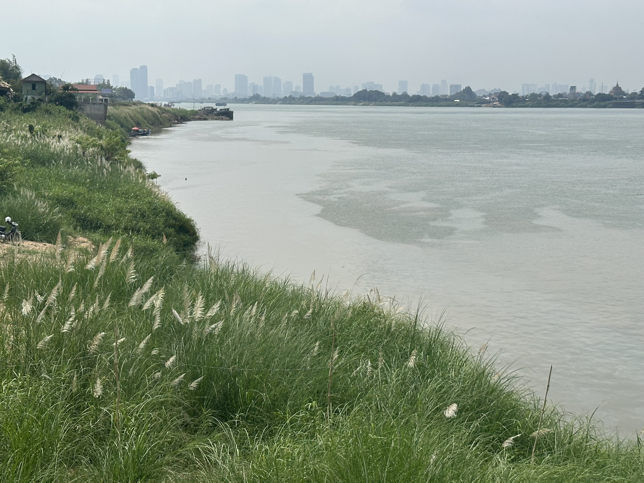 View across the Mekong River towards the Phnom Penh skyline under cloudy skies, with a grassy riverbank in the foreground.