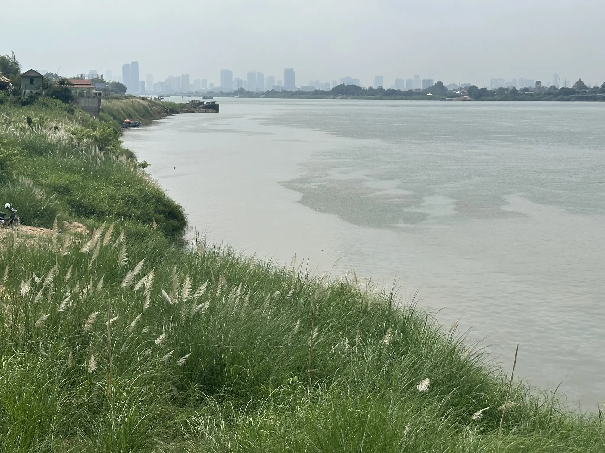 View across the Mekong River towards the Phnom Penh skyline under cloudy skies, with a grassy riverbank in the foreground.