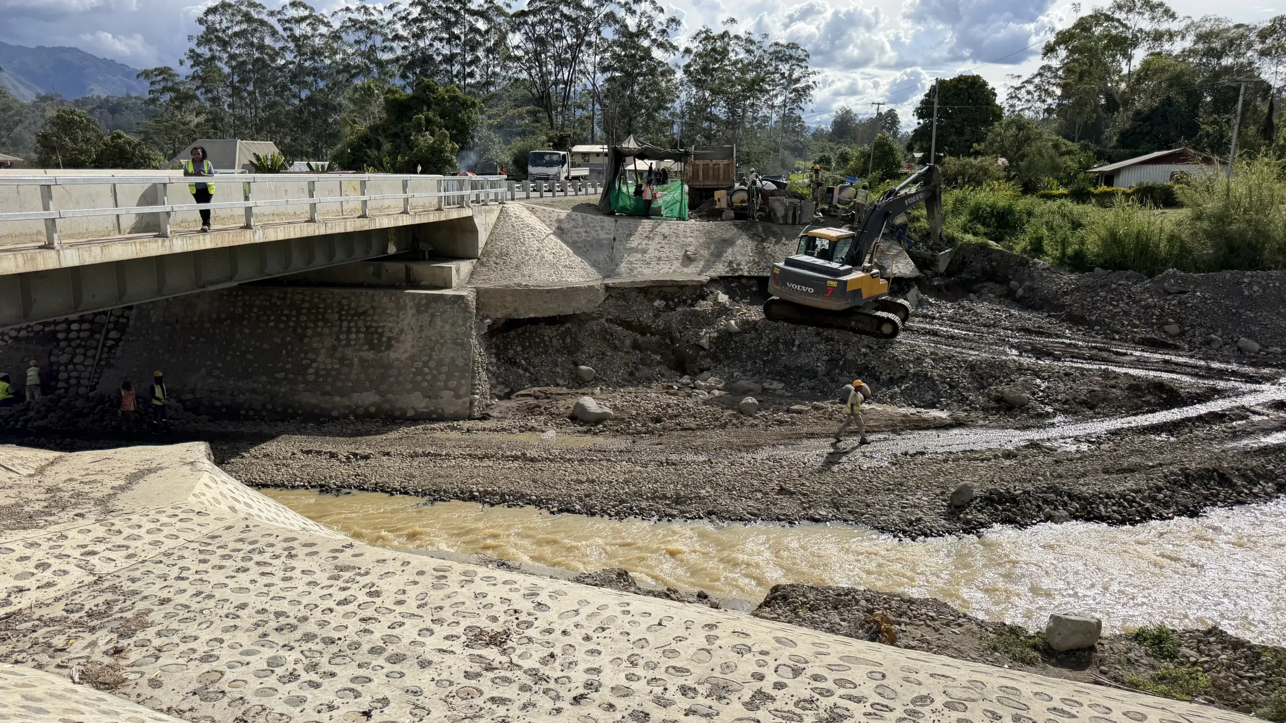 Bridge construction work over a river in Papua New Guinea, with excavators and workers on site beside the bridge structure.