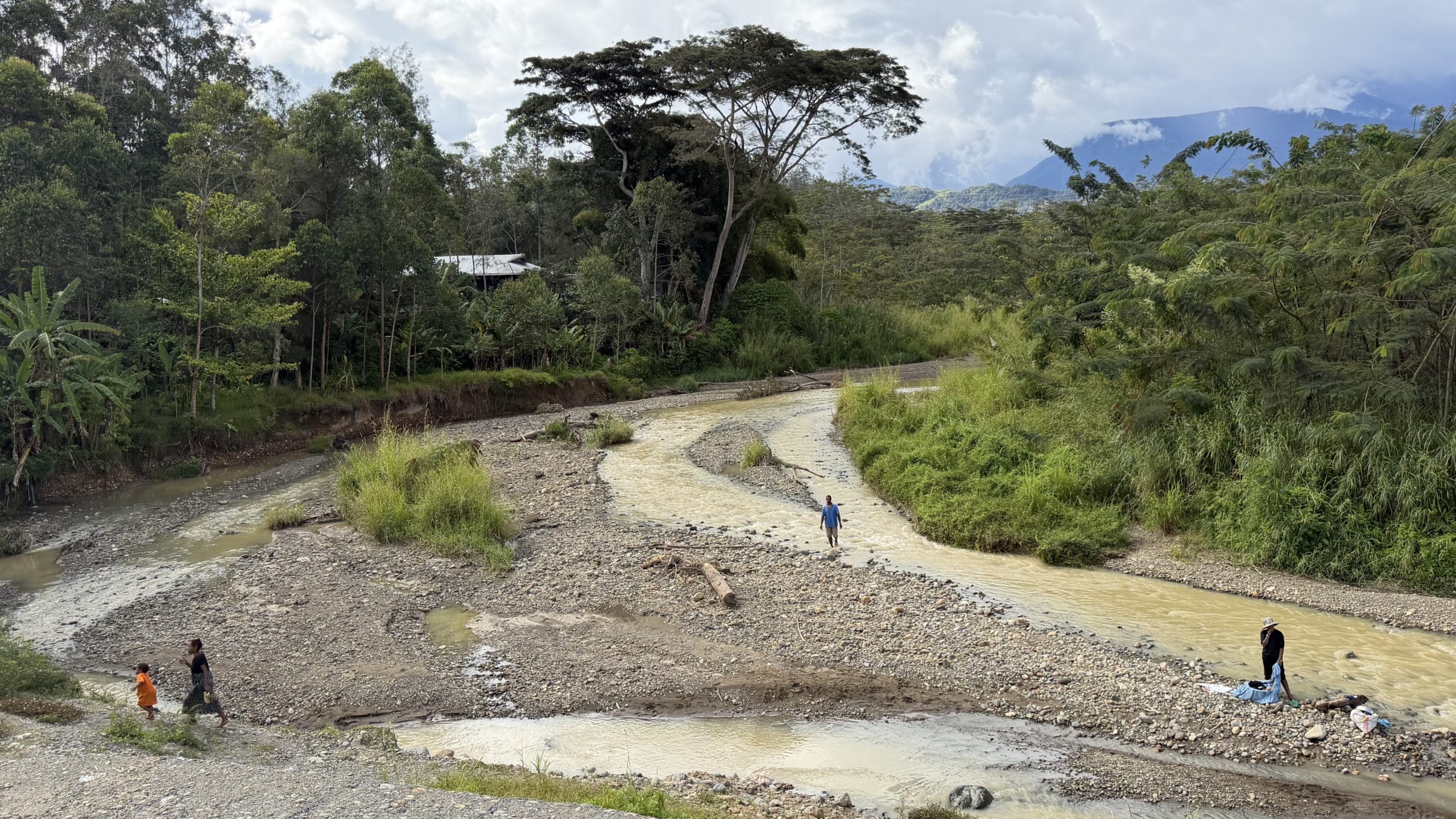 People washing clothes and crossing a shallow river in Papua New Guinea, with a gravel riverbed, dense vegetation and mountains in the background.