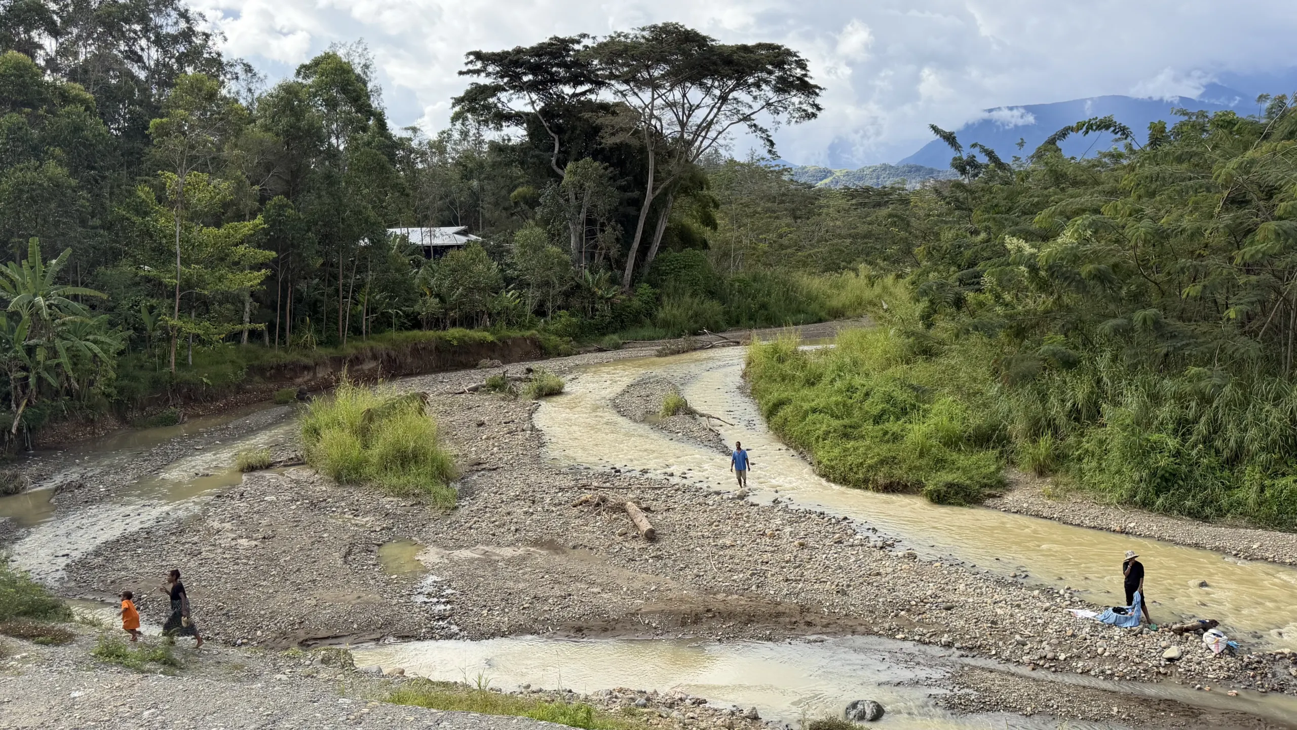 People washing clothes and crossing a shallow river in Papua New Guinea, with a gravel riverbed, dense vegetation and mountains in the background.