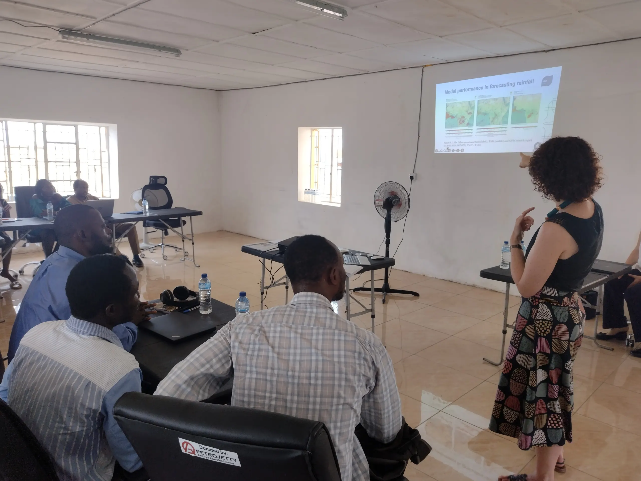 Training workshop participants seated around tables while a presenter delivers a session on rainfall forecasting and model performance using a projected presentation.