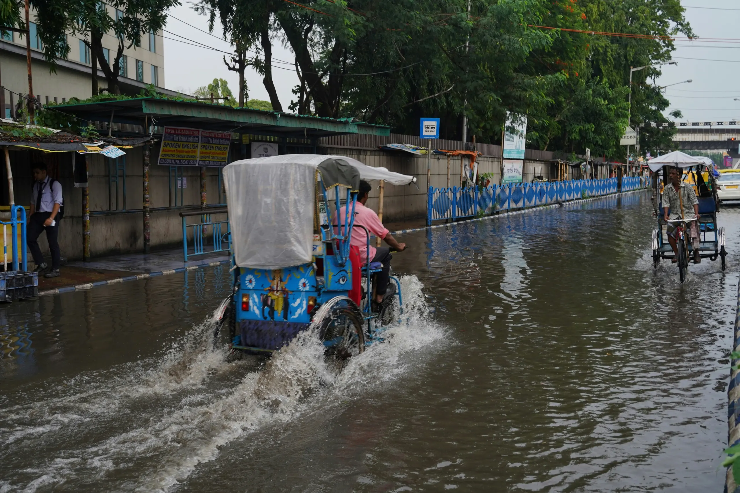 Rickshaws travelling along a flooded urban street following heavy rainfall, with standing water covering the road and buildings and trees lining the street.
